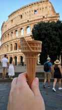 Load image into Gallery viewer, A hand holds a Conosseum Gelato and Ice Cream Cone by Gelato Supply, ingeniously crafted to mimic the iconic Colosseum, blending perfectly with the real landmark in the background as four people stroll nearby on a bright, sunny day.
